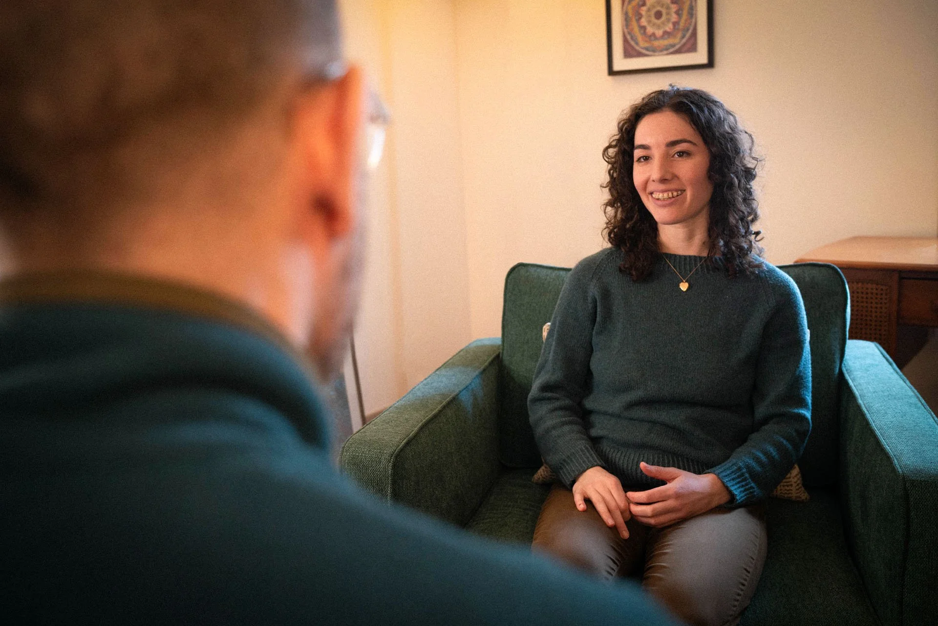 Vanessa smiling in a green armchair in her office, listening to a client seen from behind.