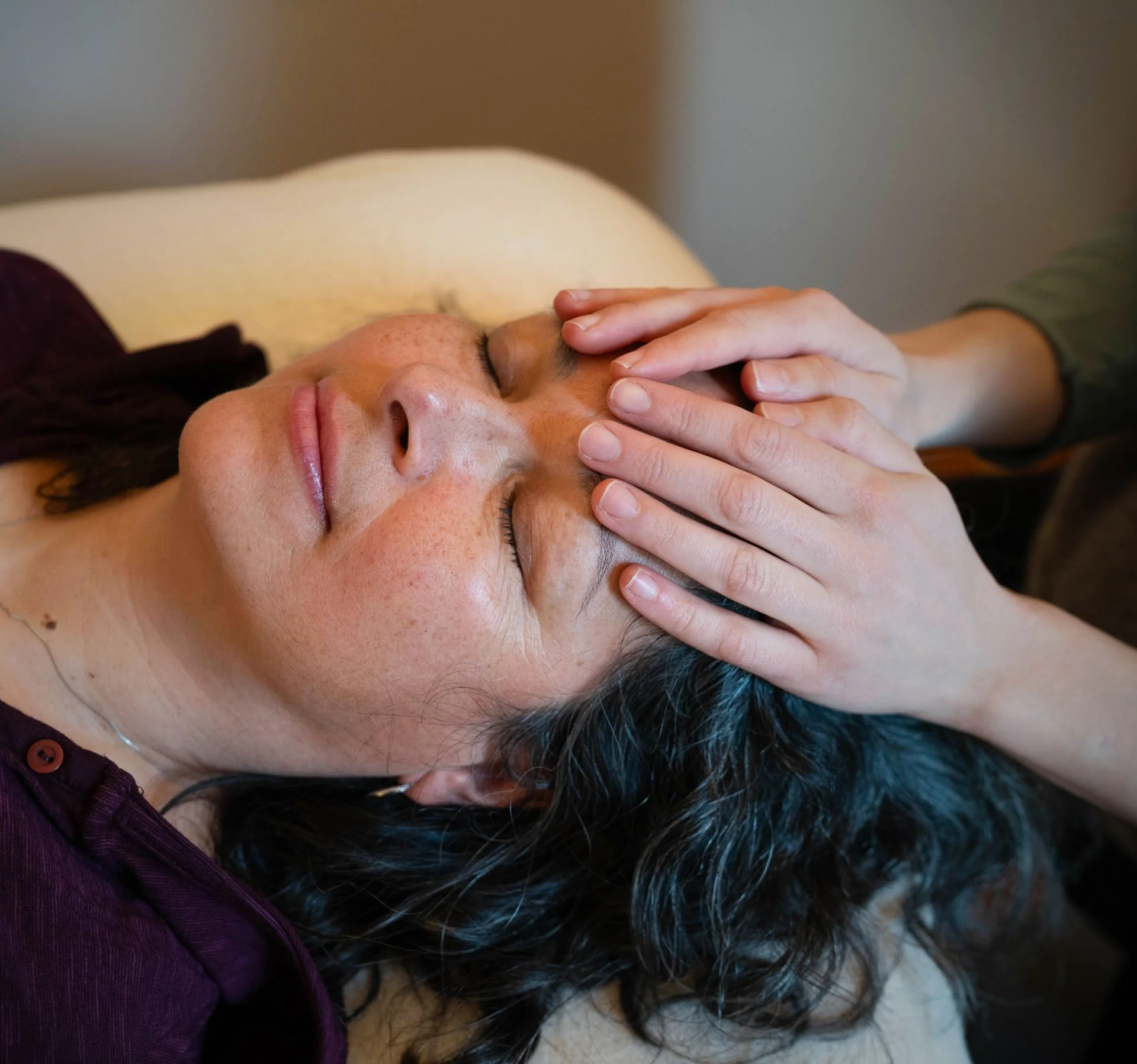 A client lying with eyes closed, Vanessa's hands resting lightly on her forehead and temple.