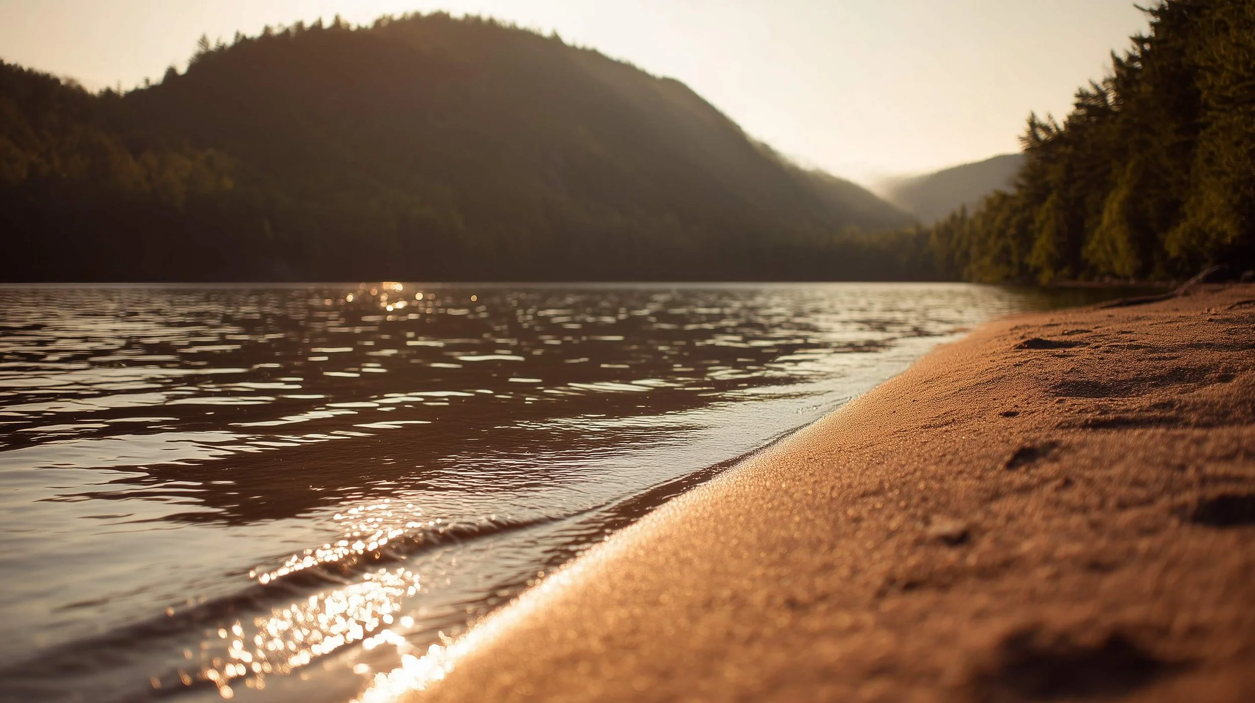 A sandy lakeshore in evening light, sun glinting on the water, with forested hills beyond.
