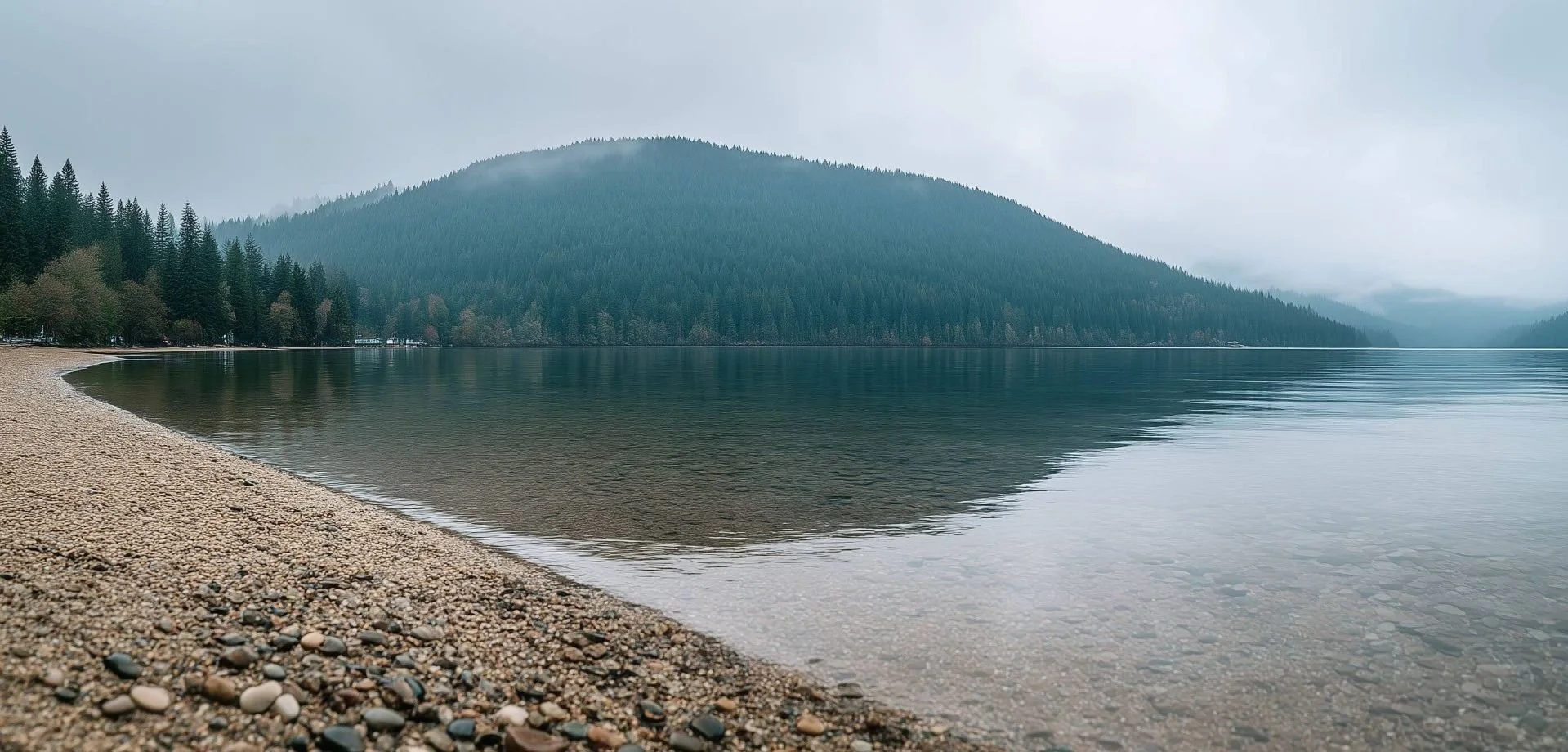 A pebble beach and a still lake under low cloud, with a forested hill on the far shore.