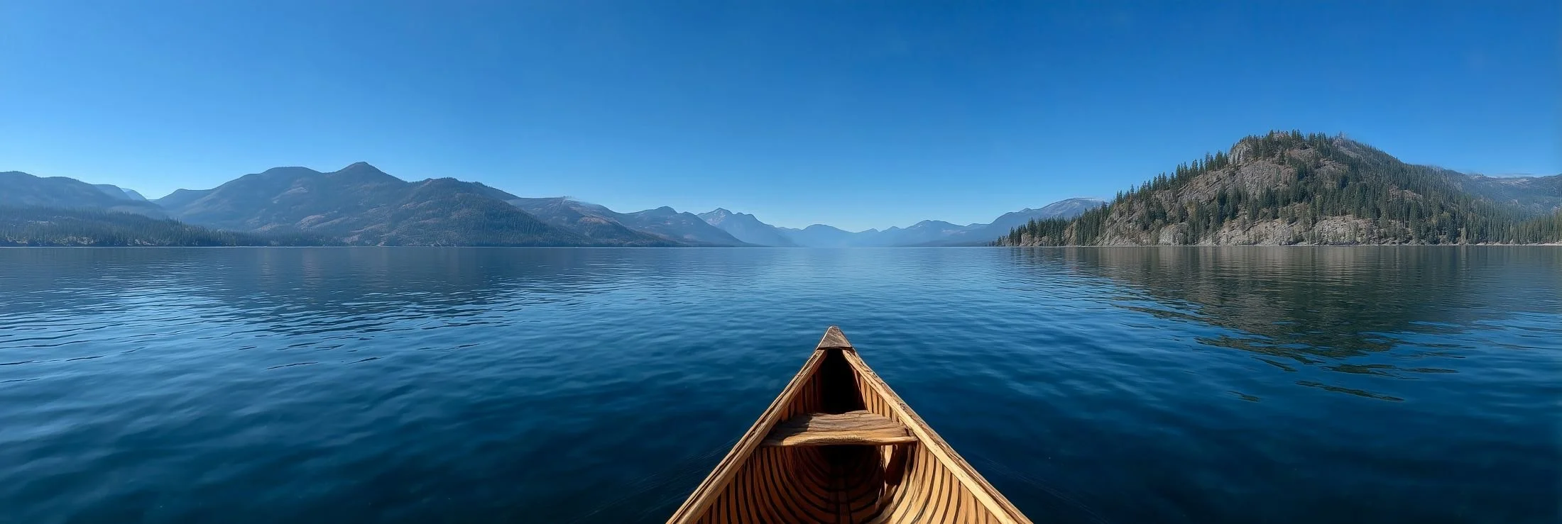 The bow of a wooden canoe on a calm blue lake, with forested mountains along both shores.