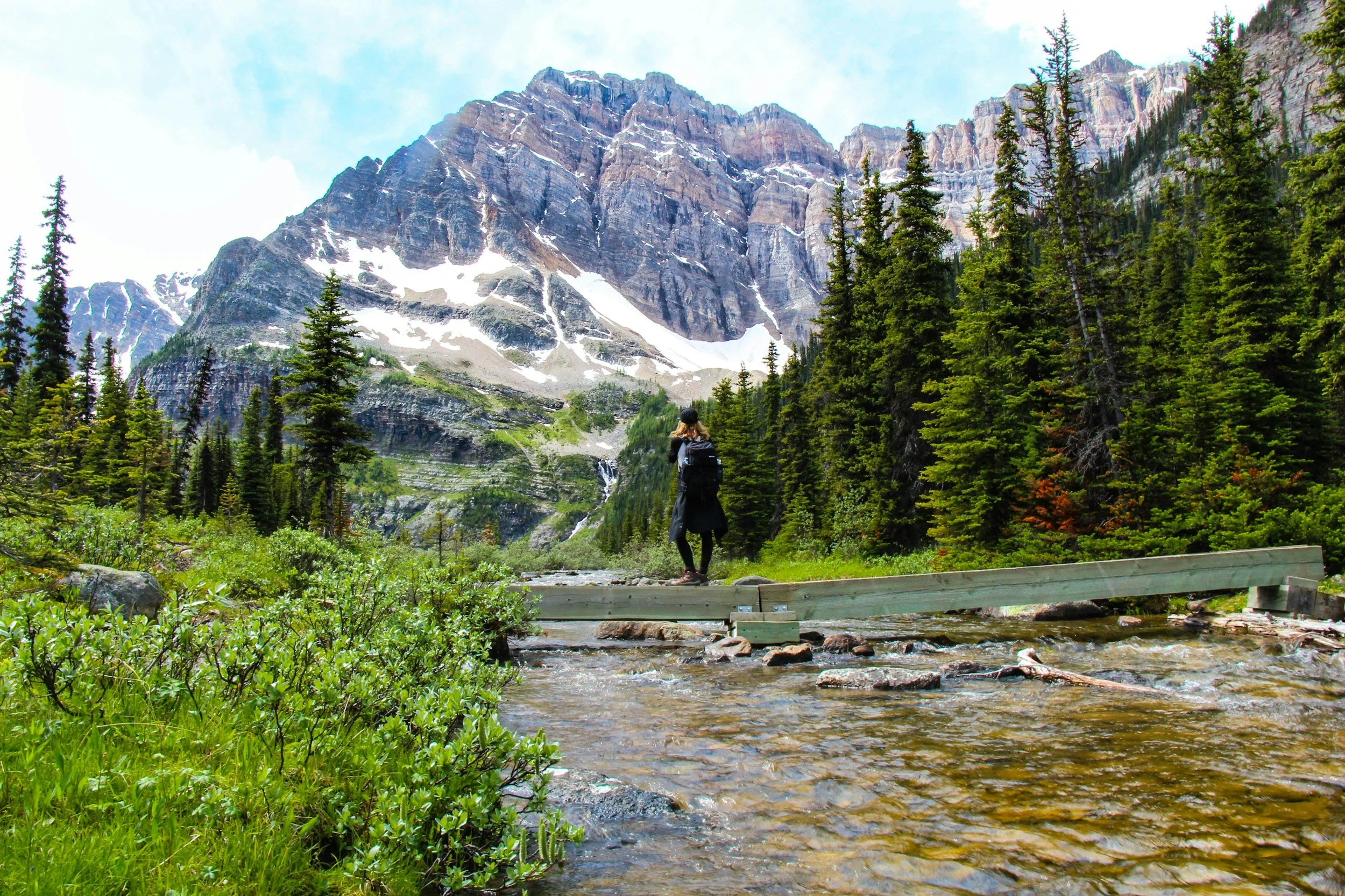 A hiker on a footbridge over a mountain stream, looking up at a snow-streaked peak.