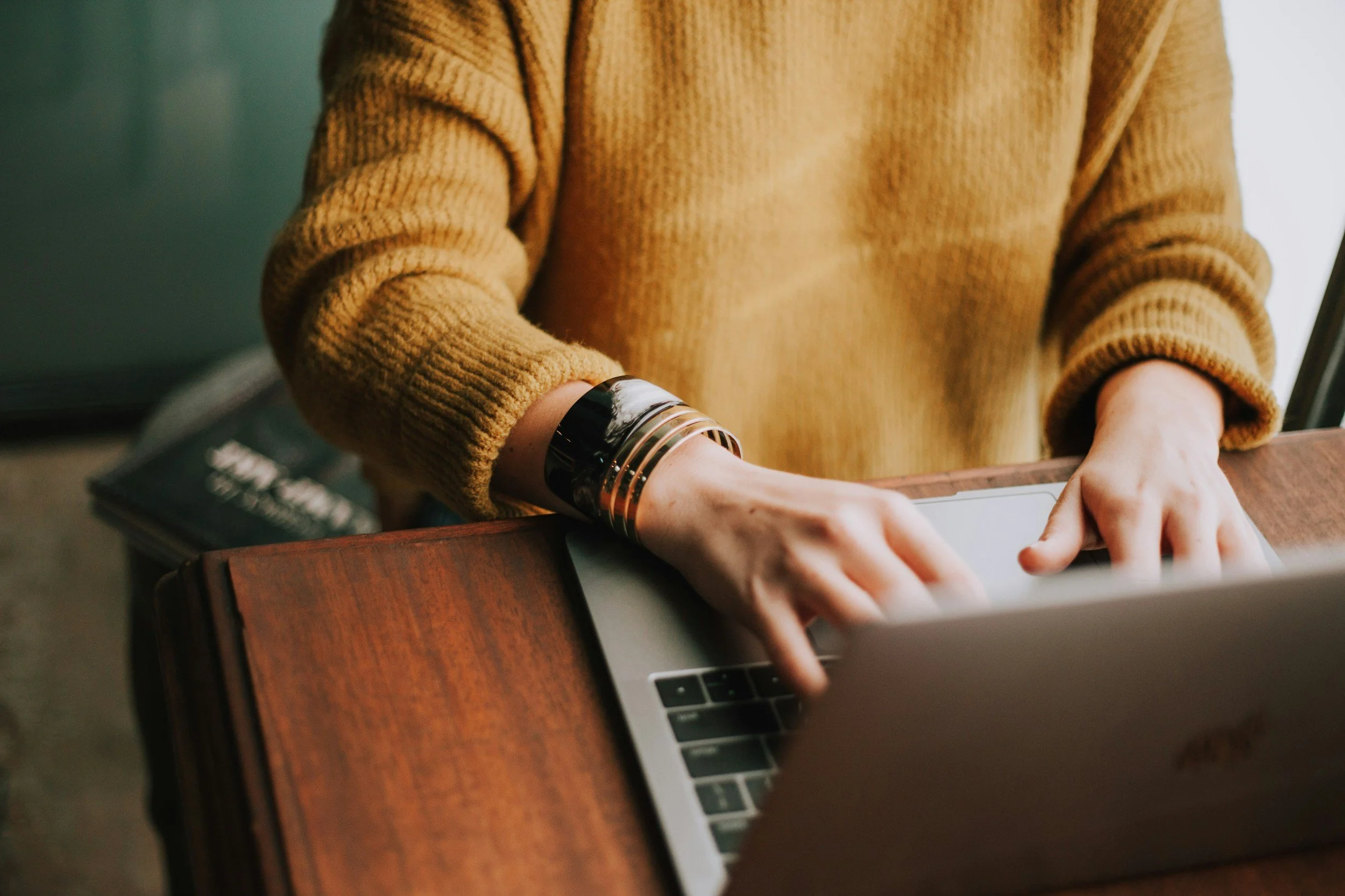 Person wearing a mustard yellow sweater typing on a silver laptop at a wooden desk.