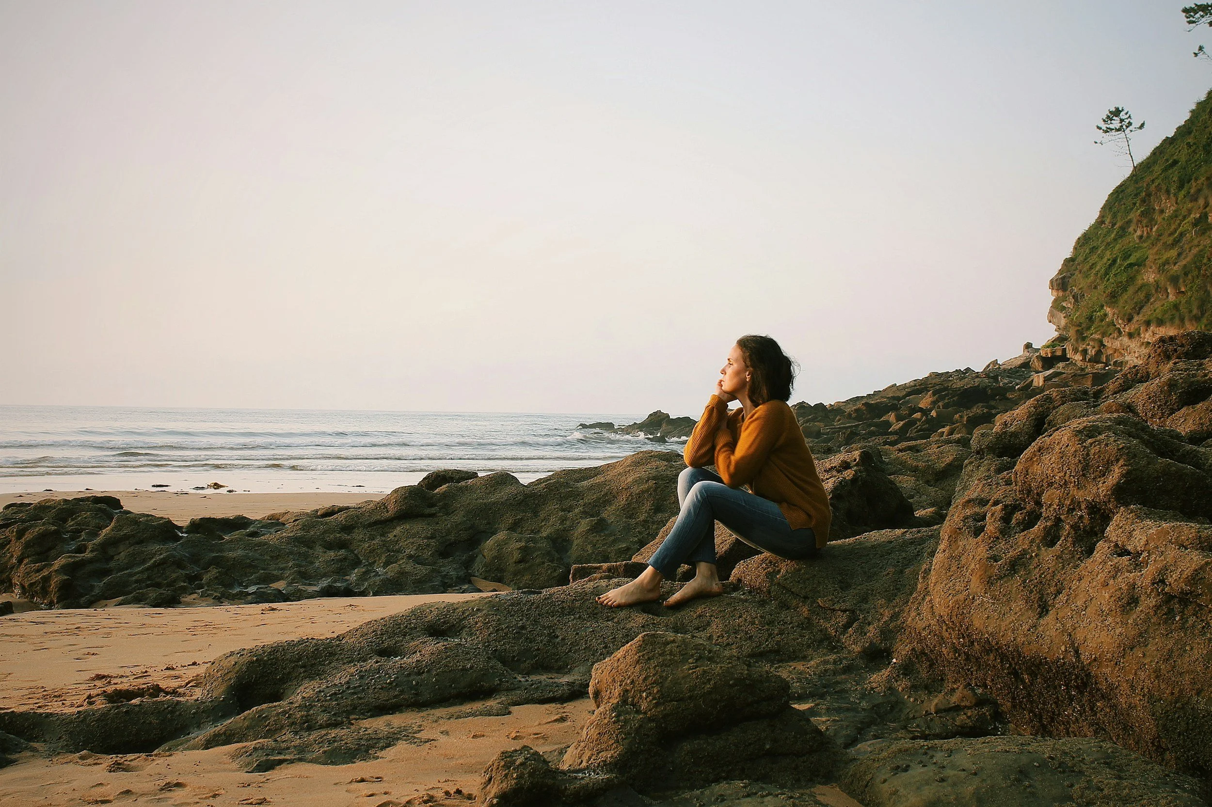 A woman sitting on rocks at the shore, chin on her hands, looking out to sea.